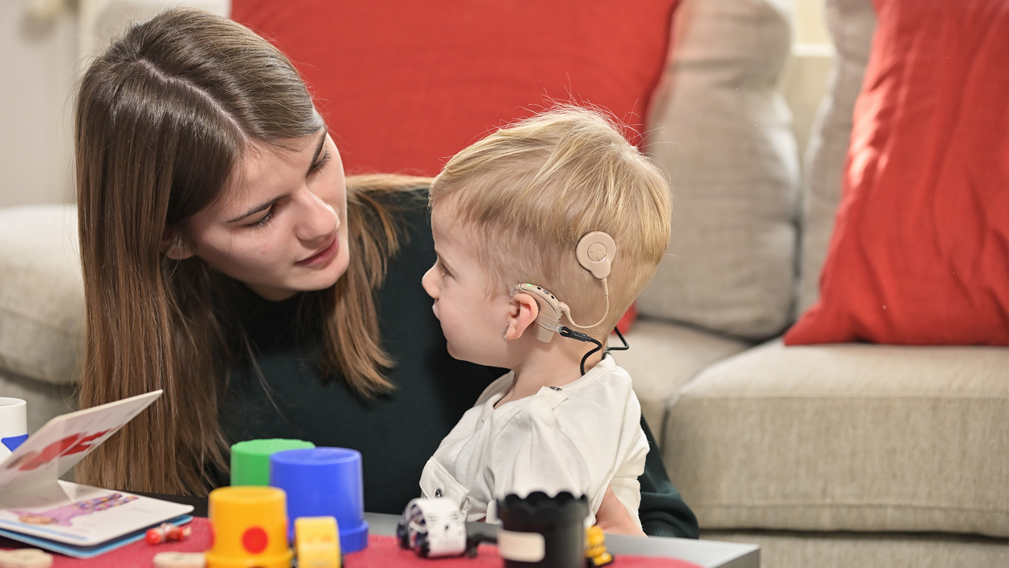 A woman sitting next to a small boy wearing a cochlear implant with building blocks on a table.