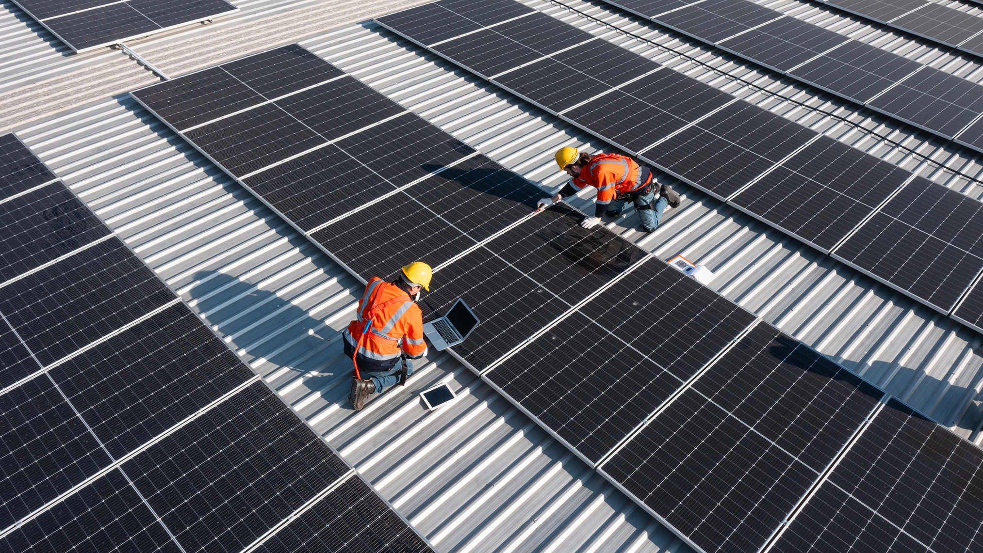 Two men kneeling on a roof monitoring fixed solar panels.